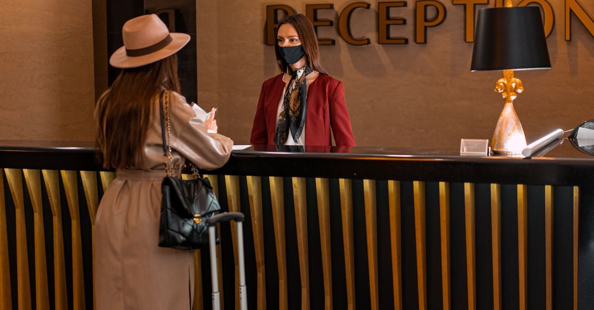 Eine Frau mit Hut und Koffer steht an einer Hotelrezeption und spricht mit einer Mitarbeiterin mit Gesichtsmaske