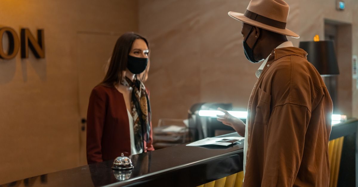 A man in a hat and a receptionist wearing face masks talk across a hotel reception desk in a professional setting