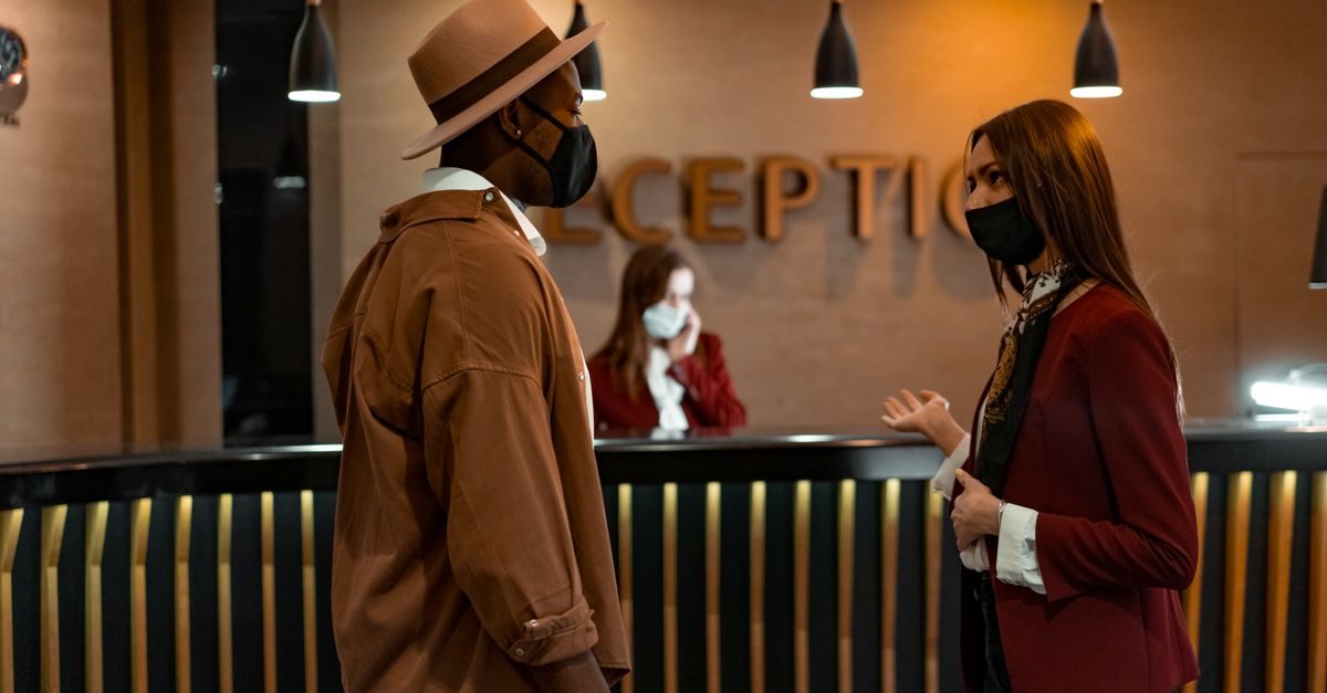 Ein Gast mit Hut und eine Hotelmitarbeiterin mit Maske unterhalten sich an einer Hotelrezeption