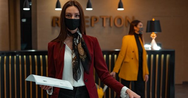 Vrouw met mondkapje en rode blazer staat bij een hotelreceptie met een collega op de achtergrond