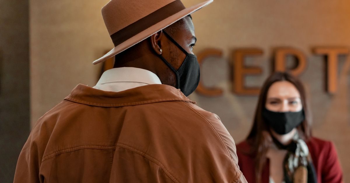 Un hombre con sombrero y mascarilla habla con una recepcionista en el mostrador de un hotel con ambiente profesional