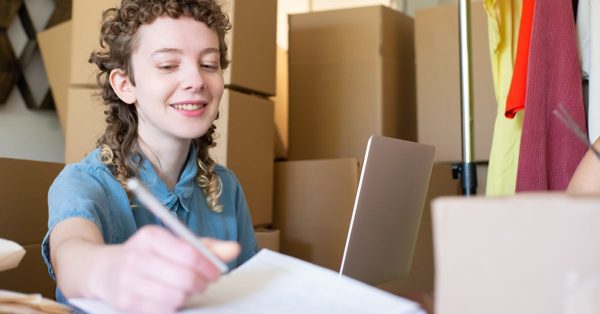 Une jeune femme souriante écrit sur un document devant son ordinateur portable au milieu de cartons de déménagement