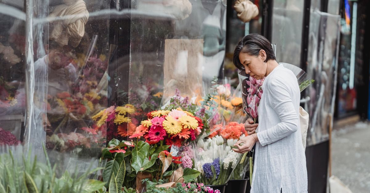 Kvinna med kort mörkt hår i ljus klänning väljer blommor utanför en blomsterbutik full av färgglada gerbera och grönska