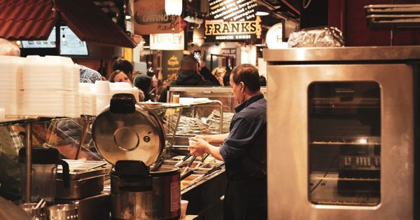 Een medewerker bereidt eten in een drukke overdekte markthal met diverse eetkraampjes op de achtergrond