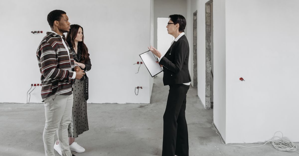 Una agente inmobiliaria con carpeta explica detalles a una pareja en un piso vacío en construcción con paredes blancas
