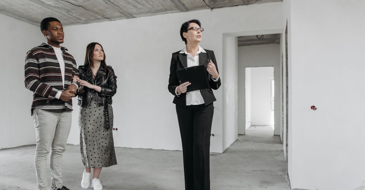 A professional estate agent shows a young couple around an unfinished, white-walled apartment during a property viewing