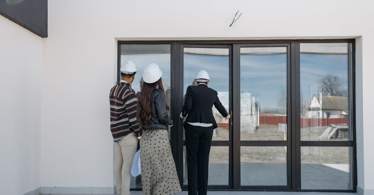 Trois personnes en casque de chantier visitent une maison neuve et ouvrent la porte vitrée donnant sur le jardin