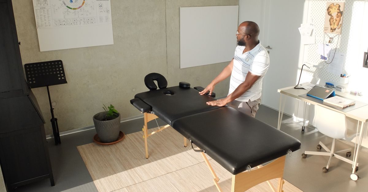 Sjukgymnast med glasögon står vid en svart massagebänk i ett ljust behandlingsrum med skrivbord och väggdekorationer