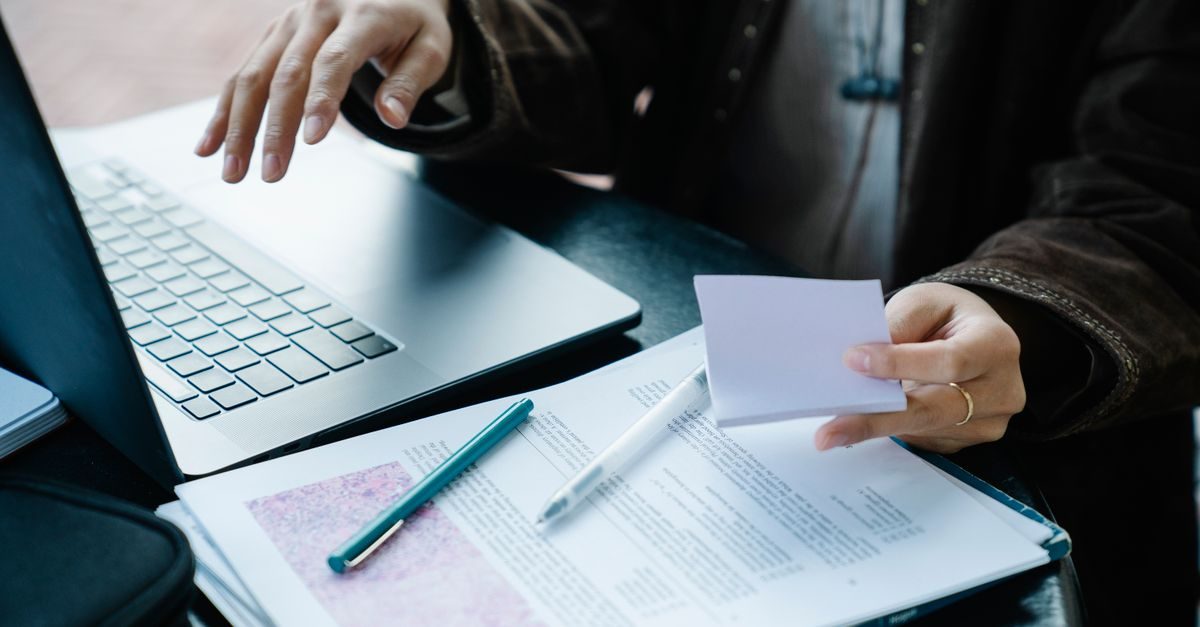 Persoon werkt aan een laptop met documenten en een leeg notitieblaadje op een bureau in een zakelijke setting