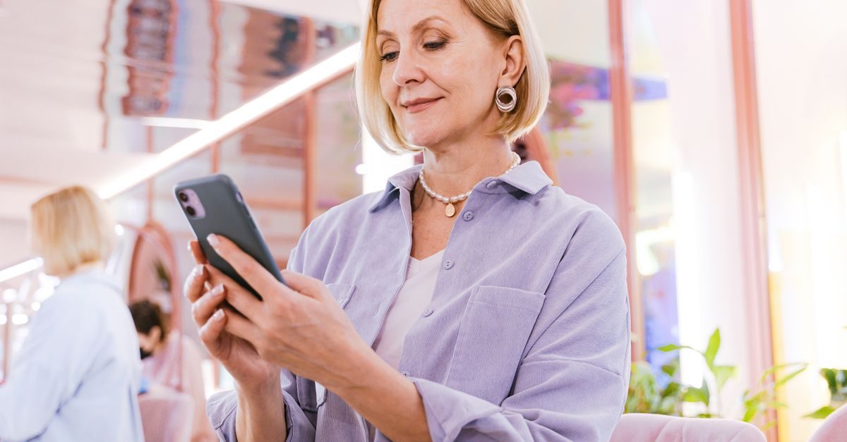A middle-aged woman with blonde hair smiles while looking at her smartphone in a bright, modern indoor setting