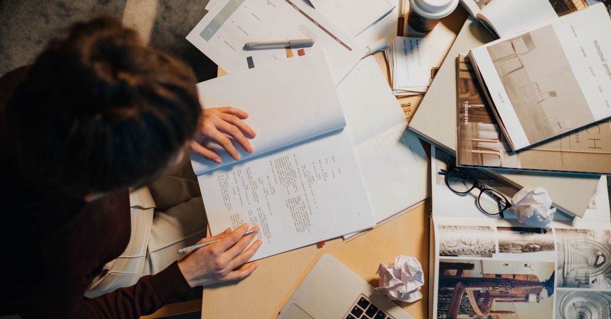 Persoon leest en schrijft aan een bureau vol met documenten, boeken, een laptop en verfrommeld papier in een studieruimte
