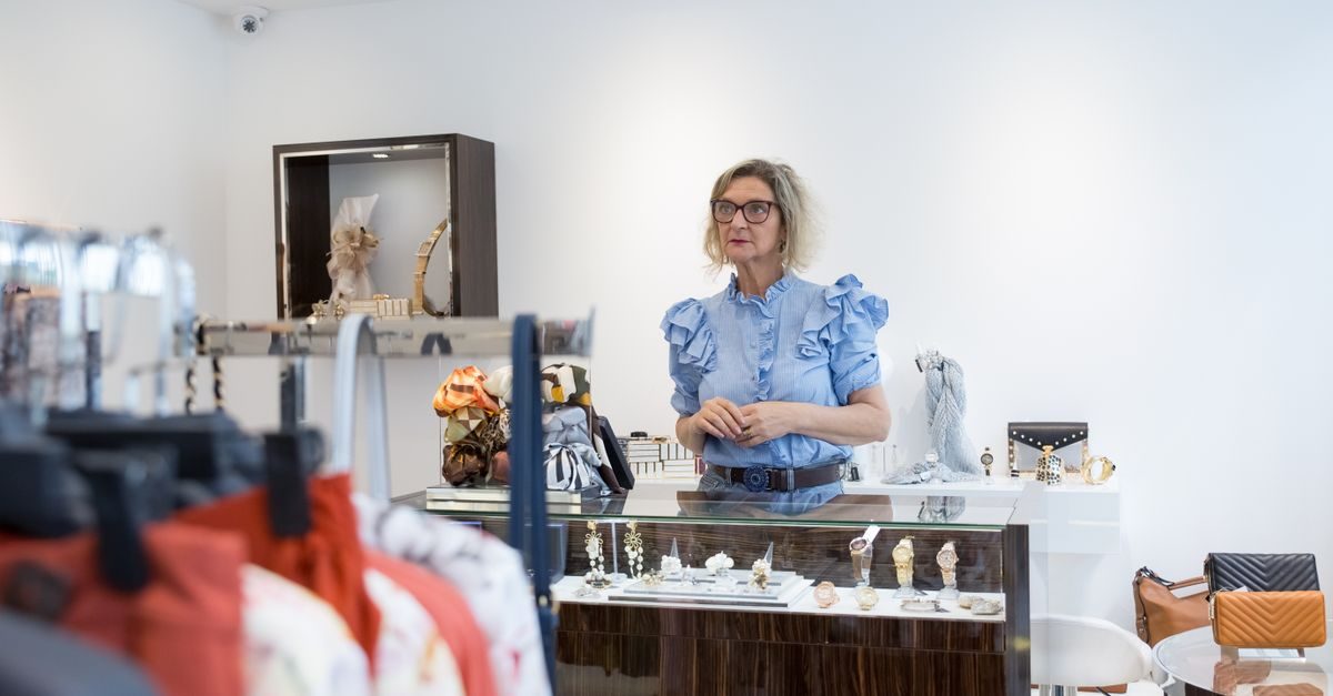 Vrouw met bril en blauwe blouse staat achter de toonbank in een kledingwinkel met accessoires en kledingrekken