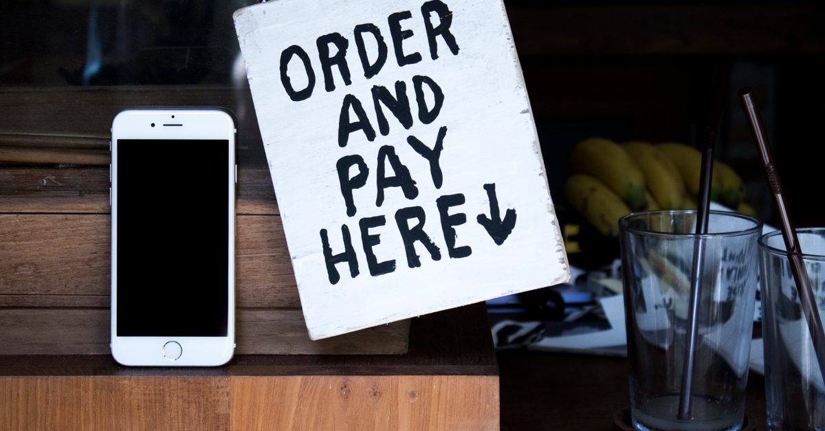 Ein weißes Smartphone steht neben einem Schild mit der Aufschrift Order and pay here auf einem Holztresen in einem Café