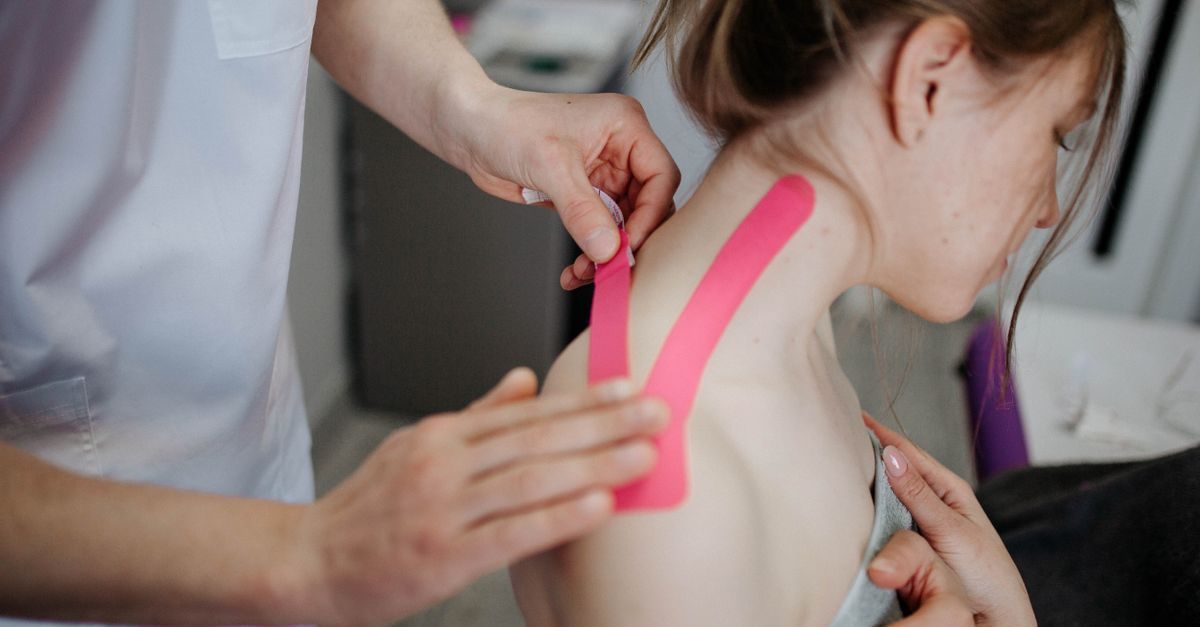 Fysiotherapeut brengt roze kinesiotape aan op de schouder en nek van een patiënt in een behandelkamer