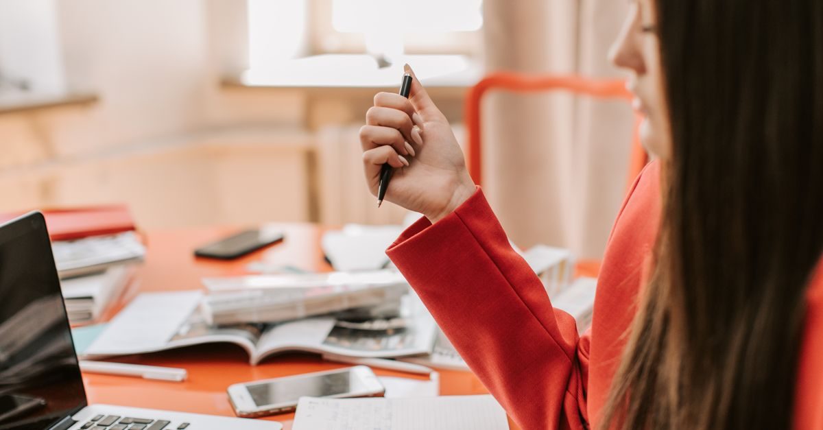 A woman in a red blazer sits at a desk with a laptop and documents, holding a pen while working in a bright office