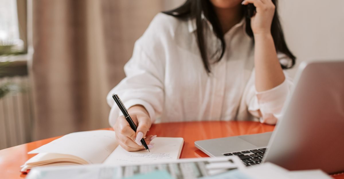 Vrouw in wit hemd telefoneert en schrijft notities in een notitieblok aan een bureau met een laptop
