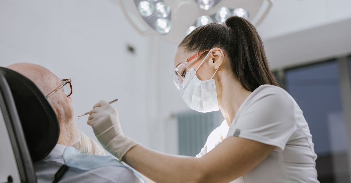 Eine Zahnärztin mit Schutzbrille und Mundschutz untersucht konzentriert einen älteren Patienten in einer modernen Praxis