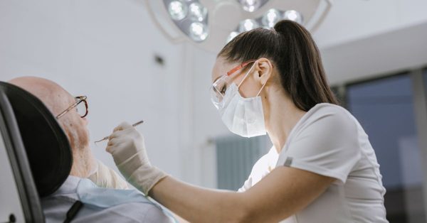 Eine Zahnärztin mit Schutzbrille und Mundschutz untersucht konzentriert einen älteren Patienten in einer modernen Praxis