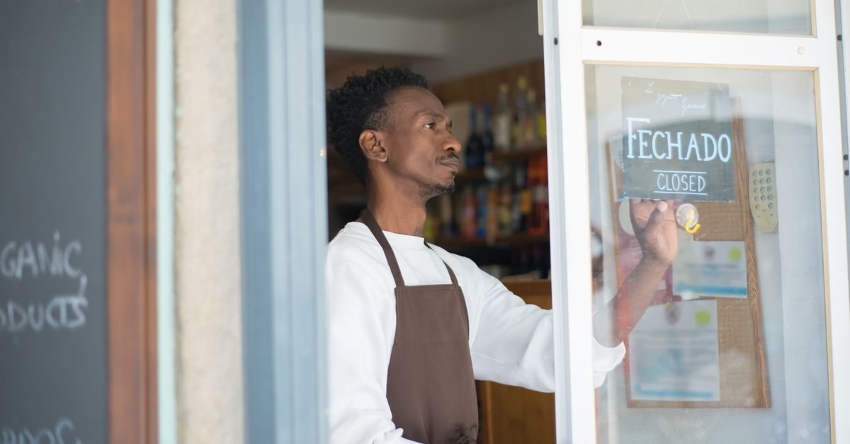 A man in a brown apron stands inside a shop, placing a closed sign on the glass door
