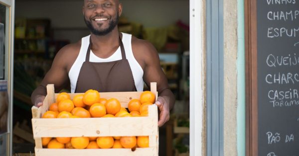 Un commerçant souriant en tablier tient une caisse d'oranges devant l'entrée de sa boutique avec un tableau noir à côté
