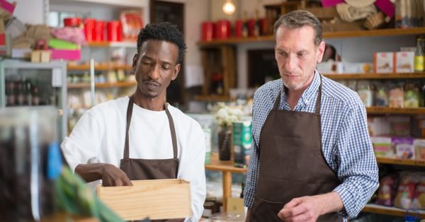 Two men wearing brown aprons work together in a grocery store, one holding a wooden box while the other looks on intently