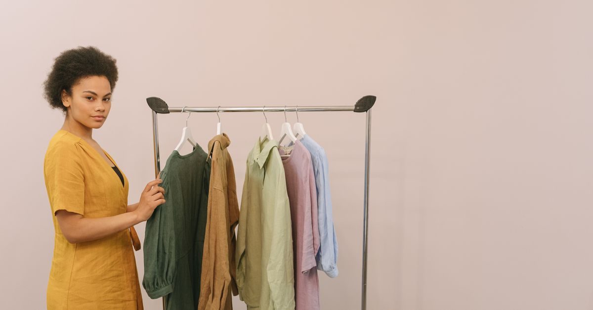A woman in a yellow dress stands beside a clothing rack with various shirts against a plain light background