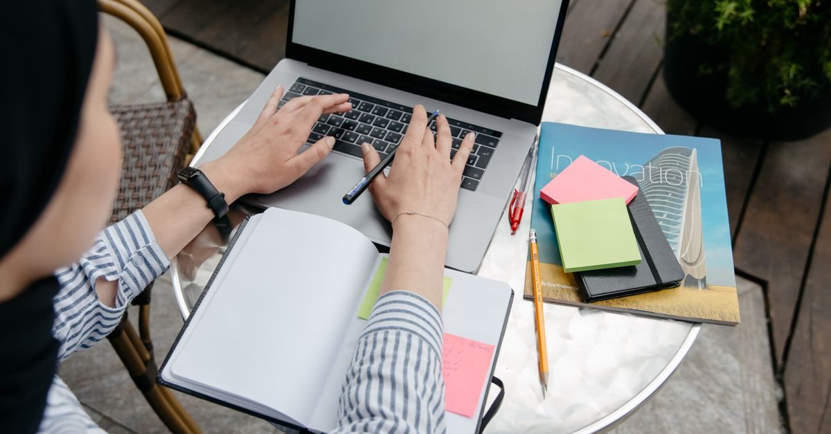 Person arbeitet an einem Laptop an einem runden Tisch mit Notizbuch, Stift und bunten Haftnotizen