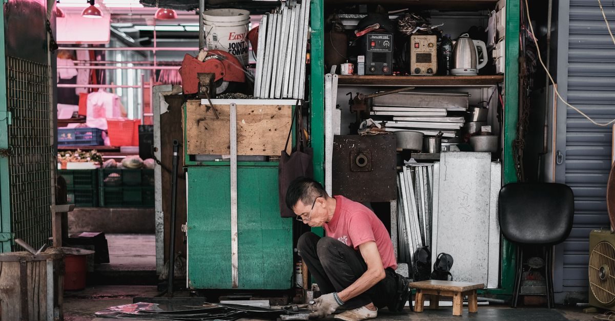 Un artisan concentré travaille accroupi dans son atelier encombré d'outils et de matériaux métalliques