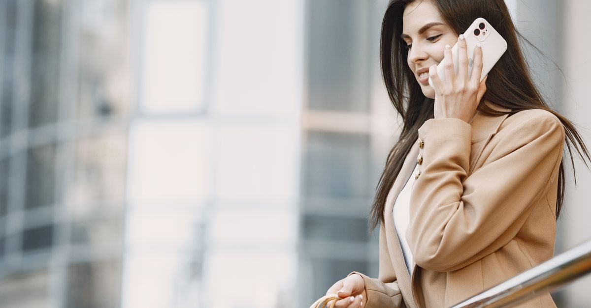 A smiling woman in a beige blazer stands outside a modern glass building while talking on her white smartphone