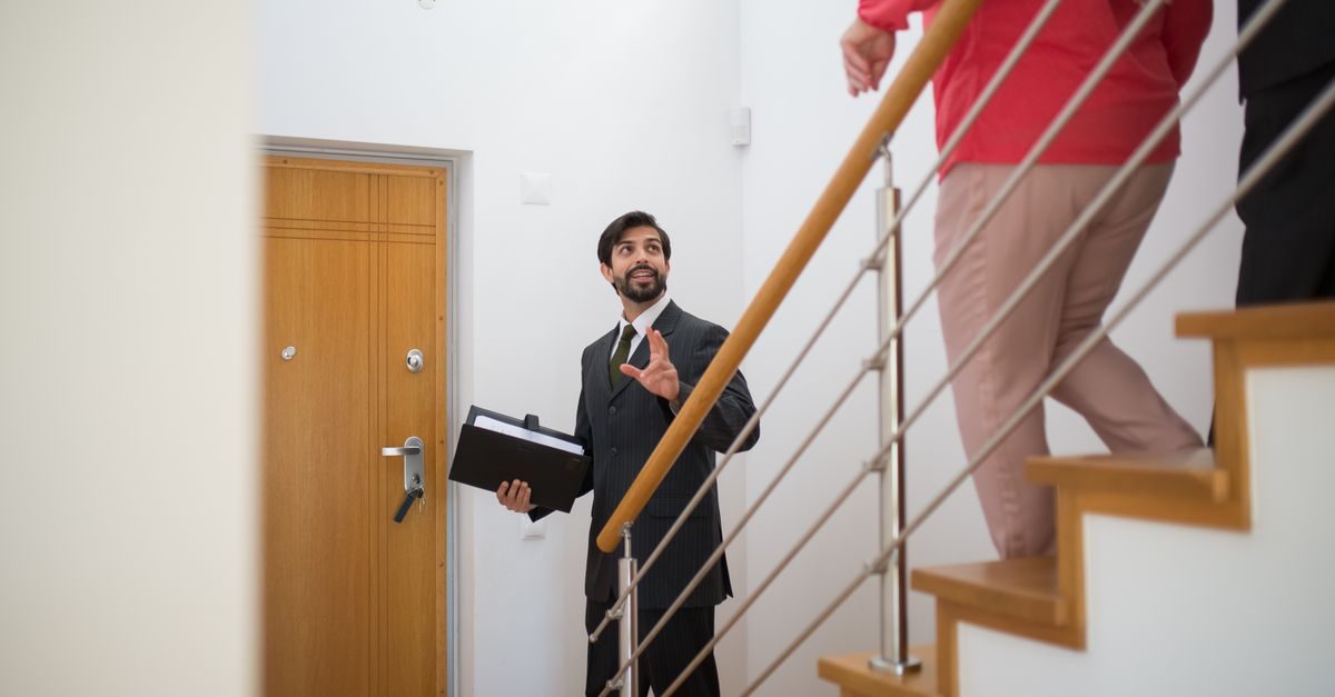 Ein Immobilienmakler im Anzug führt Kunden in einem hellen Treppenhaus durch eine Wohnungsbesichtigung