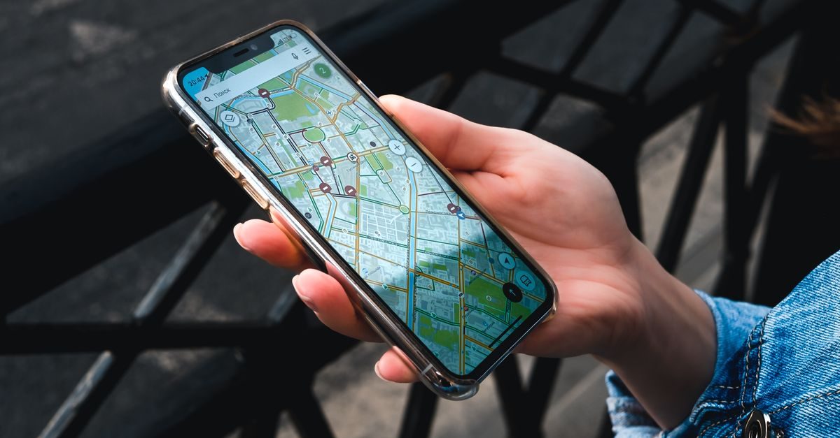 A person in a denim jacket holds a smartphone displaying a digital map interface while outdoors near a metal railing