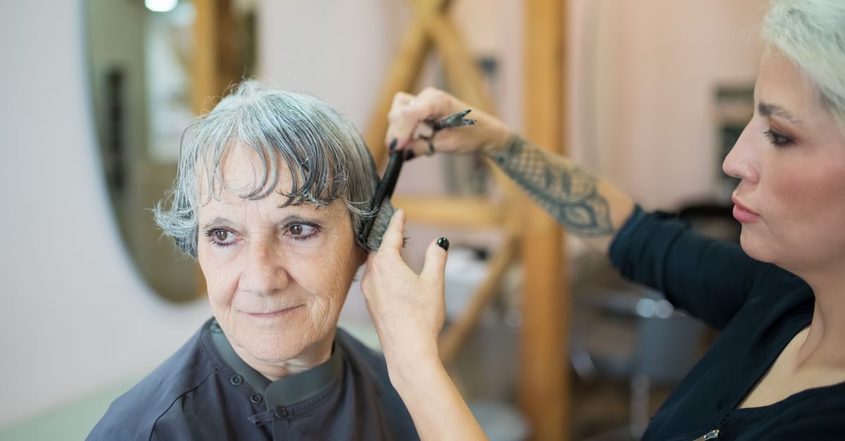 A tattooed hairdresser carefully trims the hair of an older woman in a professional salon setting