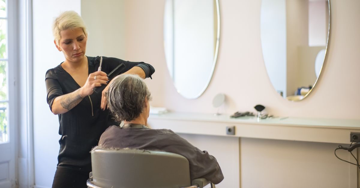 Kapper met kort blond haar knipt geconcentreerd het haar van een klant in een lichte kapsalon met ovale spiegels