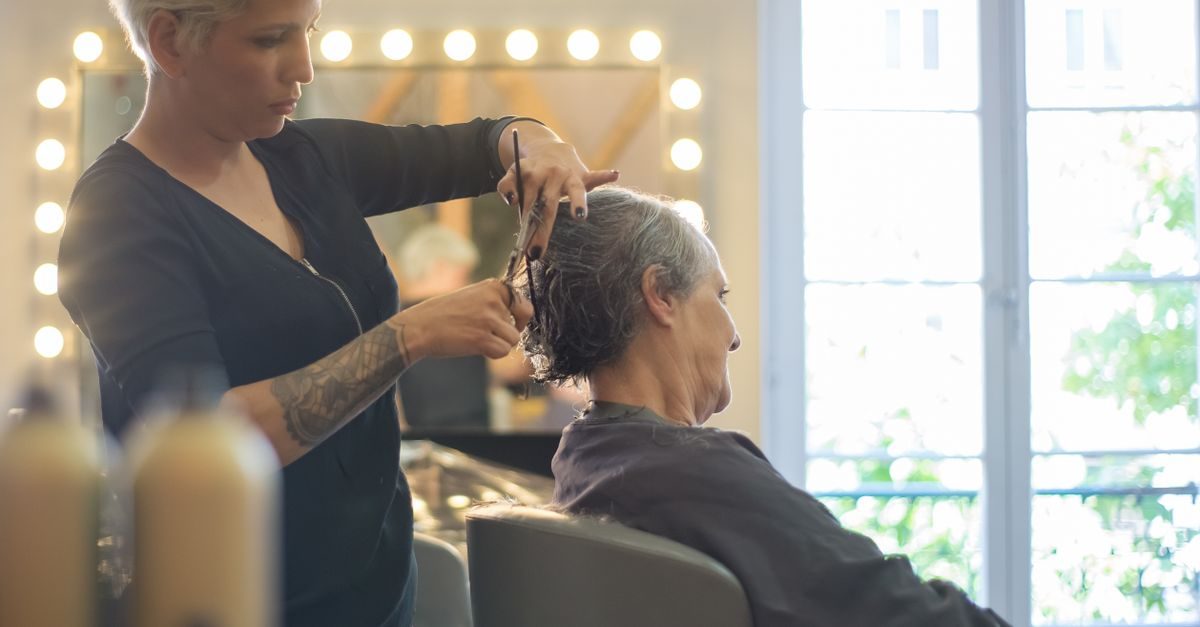 A focused hairstylist with tattoos cuts the hair of a client sitting in a bright, modern salon with vanity lights