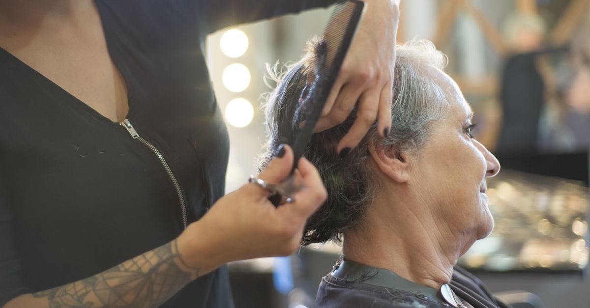 A tattooed hairdresser uses a comb and scissors to style the hair of an older woman in a professional salon setting