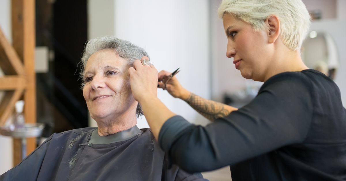 Une coiffeuse aux cheveux blonds courts coupe les cheveux d'une cliente âgée souriante dans un salon professionnel