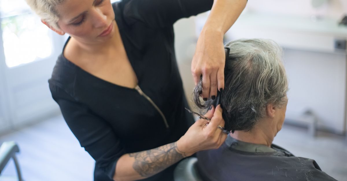 Eine Friseurin mit tätowiertem Arm schneidet konzentriert das graue Haar einer Kundin in einem hellen Salon
