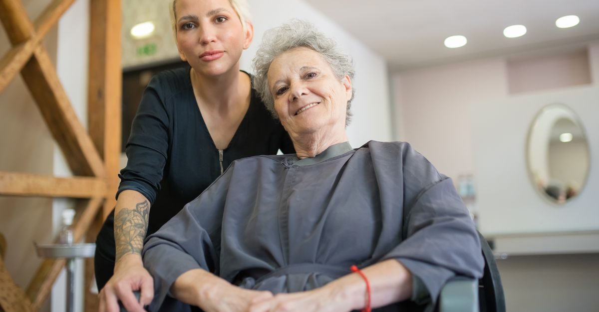 Parrucchiera bionda con tatuaggio sul braccio posa accanto a una cliente anziana sorridente seduta in un salone di bellezza