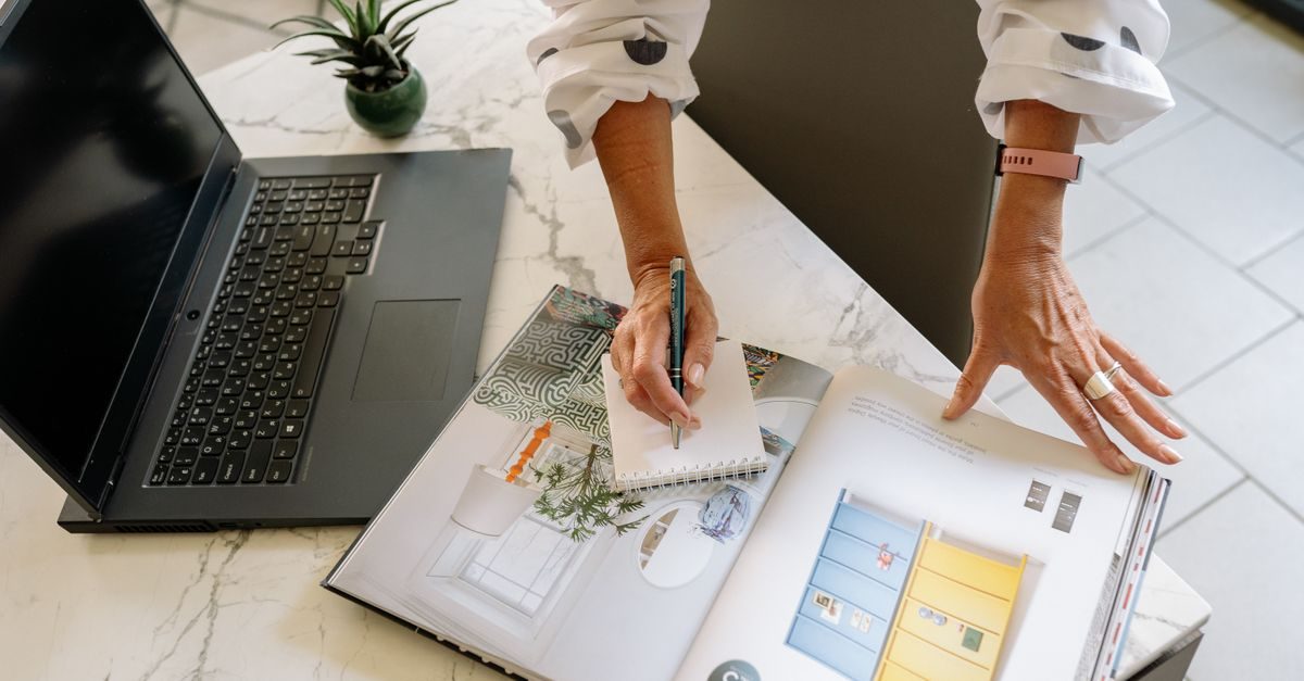A person writes in a notebook on a marble table next to an open interior design magazine and a laptop