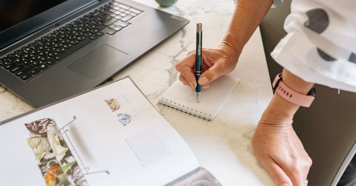 Iemand schrijft met een pen in een notitieblok naast een open boek en een laptop op een marmeren tafel