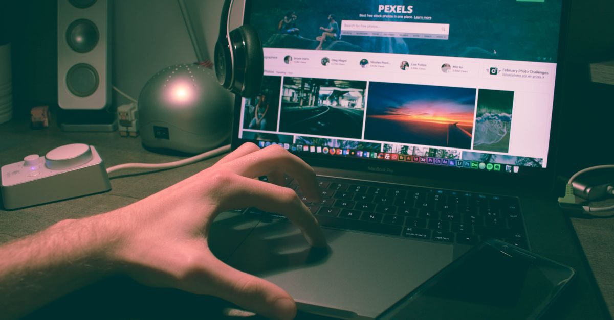 A person uses a laptop to browse the Pexels website in a dimly lit workspace with headphones and a smartphone nearby