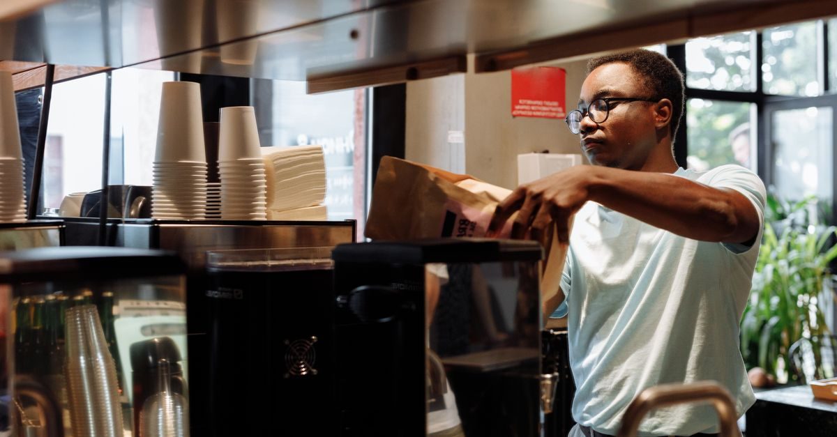 Man med glasögon i vit t-shirt packar en papperspåse bakom en kaffedisk med staplade pappersmuggar och kaffemaskin