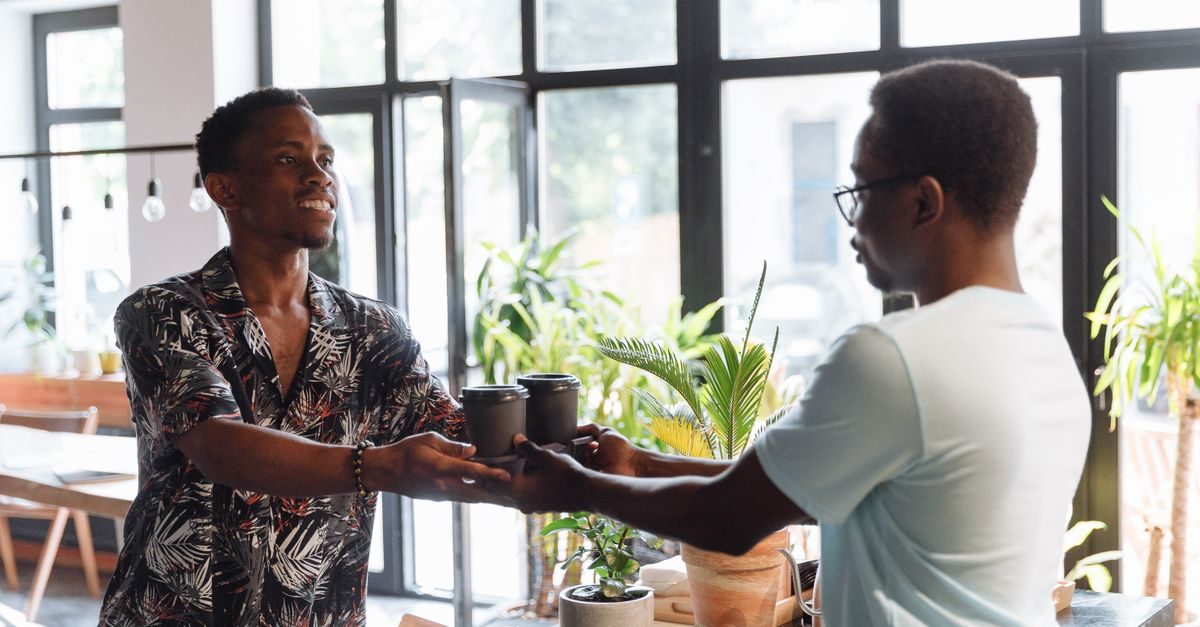 Un serveur souriant remet deux cafés à emporter à un client dans un café lumineux et végétalisé
