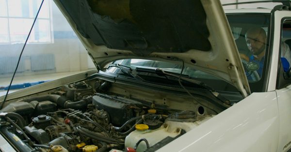 Mekaniker sitter i en bil med öppen motorhuv inne på en bilverkstad för att kontrollera motorn
