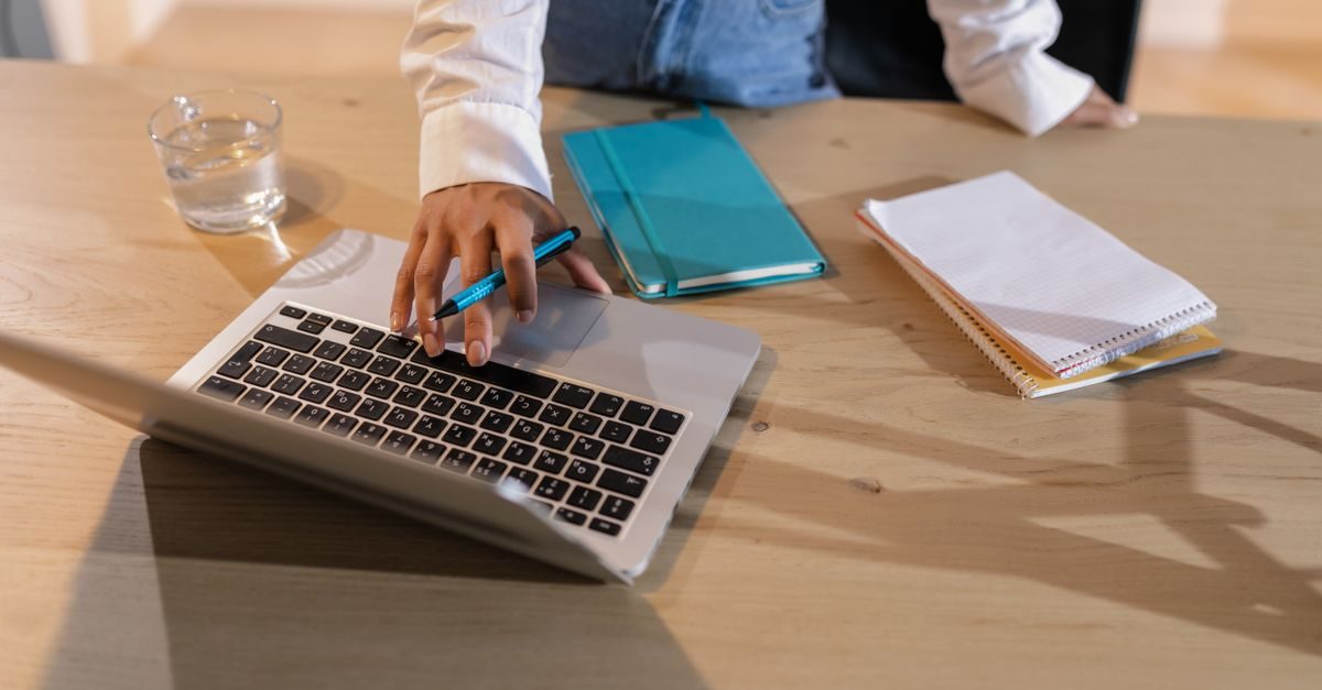 Eine Person arbeitet konzentriert an einem Laptop auf einem Holztisch mit Notizbüchern und einem Glas Wasser
