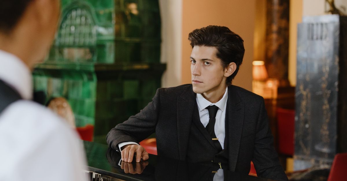 Un giovane uomo in abito elegante siede al bancone di un hotel e guarda con attenzione il receptionist di spalle