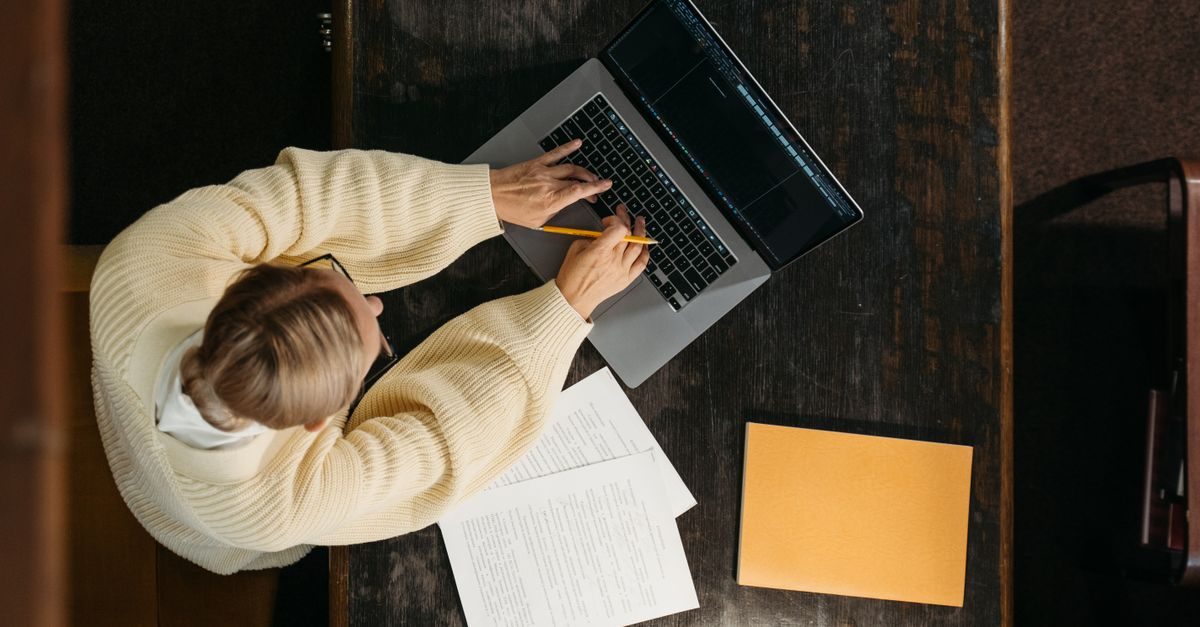 Vue de dessus d'une personne en pull beige travaillant sur un ordinateur portable avec des documents sur une table sombre