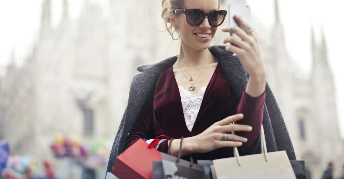 A smiling woman in a velvet dress and sunglasses holds shopping bags while looking at her smartphone outdoors