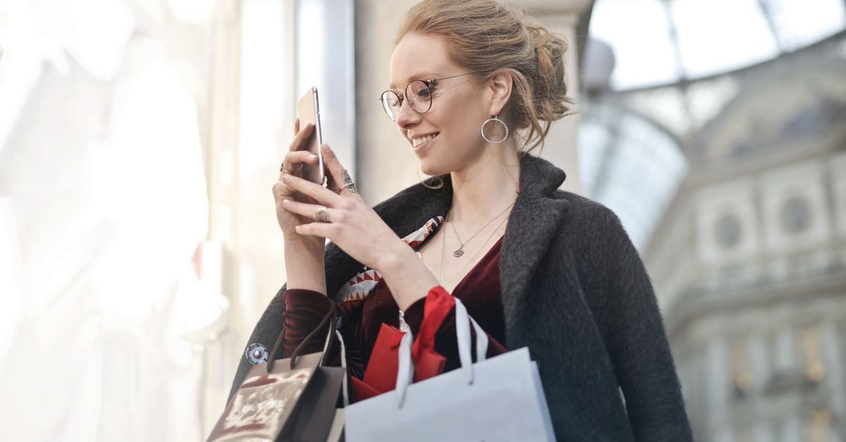 Glad kvinna med glasögon och shoppingkassar tittar i sin mobiltelefon i en ljus galleria